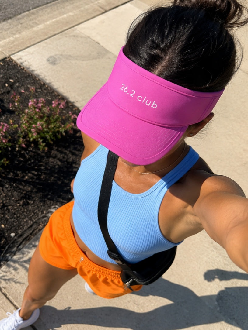 Person wearing a pink visor with '26.2 club' text, blue sports bra, and orange shorts on a sidewalk.