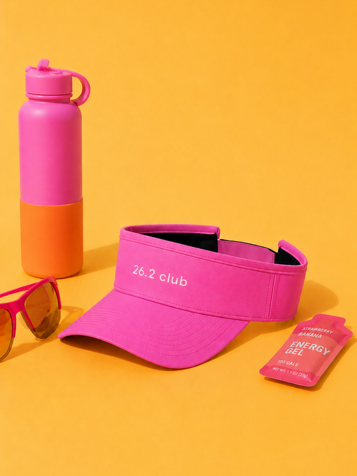Pink visor with embroidered '26.2 club' white text, pink water bottle, pink sunglasses, and pink energy gel on a yellow background.